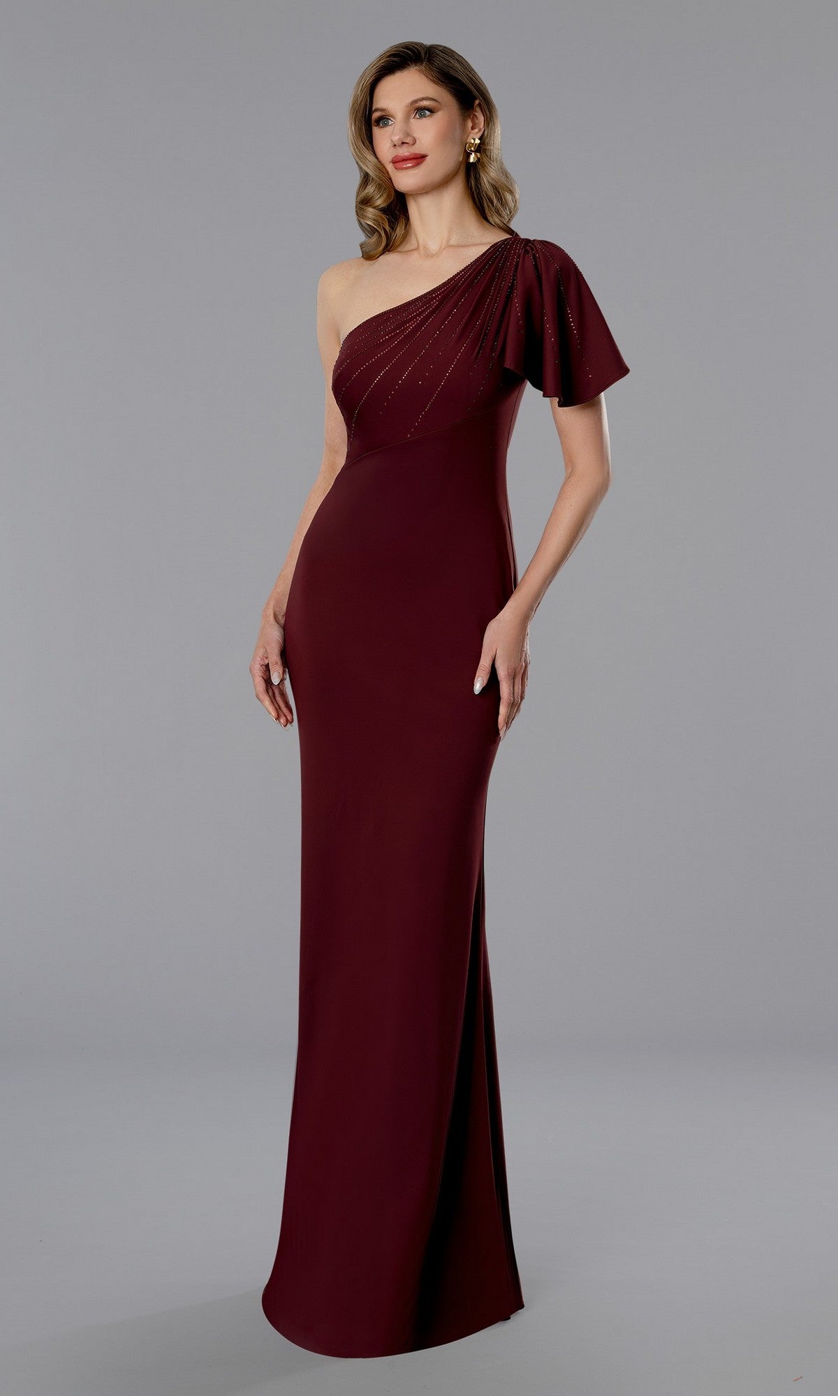 A woman stands against a plain gray background wearing the Stella Couture 25358, a long formal wine-colored dress featuring a floor-length one-shoulder design with draped sleeve and back zipper closure.